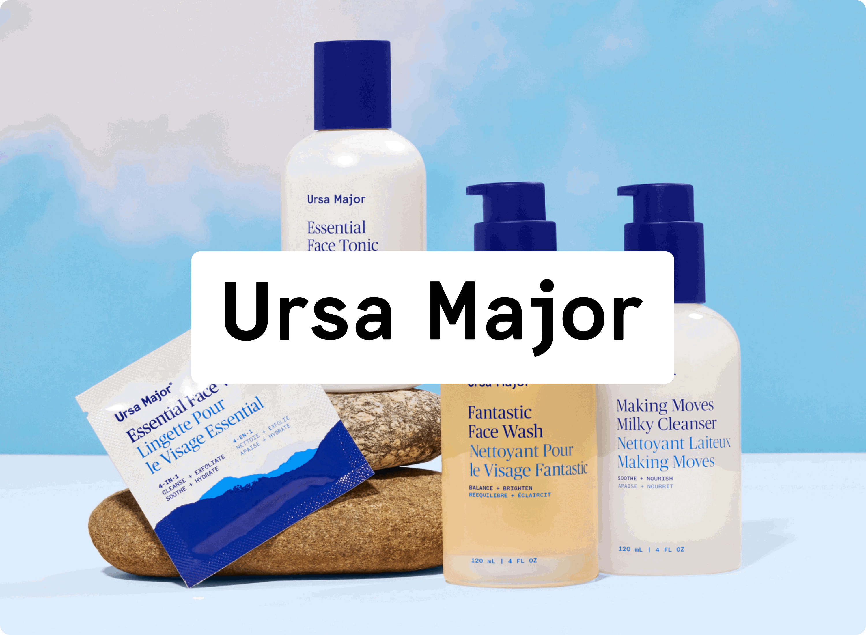 Five Ursa Major skincare products, including bottles of tonic, face wash, and cleanser, plus a cleansing wipe, are arranged on rocks against a blue sky background. A white box with Ursa Major covers part of the image.