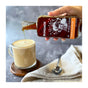A hand pours Runamok Pumpkin Spice Infused Maple Syrup into a frothy latte in a glass mug, adding cozy autumn flavors; a spoon and beige napkin sit nearby on the gray surface.