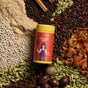 A Diaspora Spice Co. Pumpkin Spice Blend tin rests on a surface, encircled by whole spices such as cardamom pods, nutmeg, peppercorns, mace, and cinnamon sticks, along with piles of ground single-origin spices.