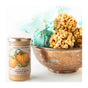 A glass jar of Savannah Bee Company Pumpkin Spice Whipped Honey, a creamy spread made with wildflower honey, sits beside a rustic brown bowl of caramel popcorn balls wrapped in blue tissue paper.