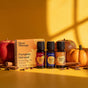Three Plant Therapy Pumpkin Harvest Essential Oil Blend Set bottles—Apple Cider, Harvest Moon, and Pumpkin Spice—are displayed with their branded box amid apples, a pumpkin, and cinnamon sticks on a yellow background, creating the perfect scene for enjoying cozy autumn aromas.