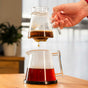 A hand lifts the Pure Over Brew Kit XL Coffee Maker with wet grounds above a carafe of brewed coffee. Fresh coffee drips into the carafe, both components of this all-glass set on a wooden table in a bright room.