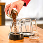 Someone pours ground coffee from a small glass jar into the Pure Over Brew Kit XL Coffee Maker, featuring a metal handle, placed on a wooden table.