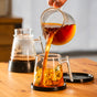 A hand pours hot coffee from the Pure Over Brew Kit XL Coffee Maker into a clear cup on a wooden table. Another carafe with coffee grounds sits in the background, and a black lid rests beside this plastic free brewer.