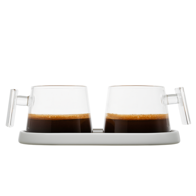 The Pure Over Espresso Cup Set features two glass cups with clear handles, filled with dark coffee and rich crema, placed side by side on a white tray against a plain white background.