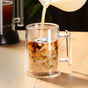 Milk is being poured into a Pure Over Double-Walled Mug Set (Set of 2), filled with iced coffee on a light wooden surface, creating swirling patterns as the liquids mix.