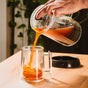 A hand pours tea from a glass French press into a Pure Over Double-Walled Mug (Set of 2) on a wooden table, with green plants softly blurred in the background.