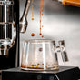 Fresh espresso pours from the machine’s portafilter into a Pure Over Espresso Cup (3.5 oz, Set of 2), displaying rich crema and droplets in the clear glass. The espresso machine is partially visible in the background.