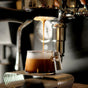 Espresso pours from a coffee machine into a Pure Over Espresso Cup (3.5 oz), Set of 2, forming a rich crema on top. The warmly lit scene highlights the inviting atmosphere and showcases the beauty of quality espresso preparation.