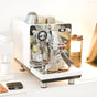 A white espresso machine with a reflective front panel brews two shots of espresso with rich crema into Pure Over Espresso Cups (3.5 oz), Set of 2, while several cups rest on top and coffee accessories sit nearby.