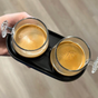 A hand holds a black tray with two Pure Over Espresso Cup Set glasses filled with fresh espresso, each topped with rich crema. A blurred, light-colored floor appears in the background.