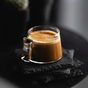 The Pure Over Espresso Cup Set displays rich espresso topped with thick crema, served in clear glass cups and placed on stacked black slate coasters against a dark, blurred background.