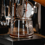 Close-up of rich espresso pouring from a machine into the Pure Over Espresso Measuring Cup, with a gauge in the background and the cup resting on a black platform—ideal for any barista.