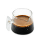The Pure Over Espresso Measuring Cup, made of clear borosilicate glass, is filled with espresso that shows off a rich crema layer on top, set against a white background.