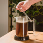 A hand stirs coffee grounds in the Pure Over French Press Coffee Maker made of borosilicate glass on a wooden table, with green plants in the background, setting the scene for brewing rich, full-bodied coffee.
