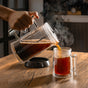 A hand pours hot, full-bodied coffee from the Pure Over French Press Coffee Maker into a clear borosilicate glass mug on a wooden table, with steam rising and a softly blurred indoor background.