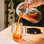 A hand pours coffee from the Pure Over French Press Coffee Maker into a clear double-walled mug on a wooden table, with the black French press lid nearby and green plants in the background.