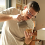 A man with glasses pours steamed milk from a silver pitcher into a Pure Over Cappuccino Cup (6.5 oz), crafting latte art in a sunlit room decorated with framed pictures.