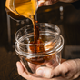 A close-up of a hand pouring coffee from a glass container into a clear, double-walled Latte Cup by Pure Over against a dark background.