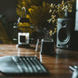 A steaming Signature Mug (14 oz) by Pure Over sits on a wooden desk alongside a keyboard, monitor, speaker, lantern candle, and potted plants in the background, creating a cozy and inviting workspace.