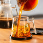 Hot coffee pours from a glass carafe into the Pure Over Signature Mug (14 oz) on a wooden table. Another carafe sits in the background as steam and coffee swirls rise inside this toxin-free borosilicate glass mug.
