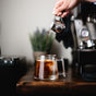 A hand pours coffee from a metal pitcher into the 14 oz Pure Over Signature Mug, made of borosilicate glass and filled with ice on a wooden table; ceramic coaster, coffee machine, and plant are blurred in the background.