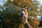 Outdoors, someone sprays a small dark bottle of Ursa Major’s Forest Fix Spray Deodorant, releasing a soft mist, with blurred autumn trees in the background.