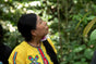 A woman with long dark hair, dressed in a bright yellow embroidered shirt and pink flower earrings, gazes upward amidst a lush green forest—an inspiring image of Indigenous-led conservation work echoing the dedication behind Grove Co.’s Forest and Biodiversity Protection initiative.