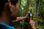 A person uses a Grove Co. Forest and Biodiversity Protection handheld GPS device against a tree wrapped with yellow tape in a forest, documenting its location as part of Indigenous-led conservation efforts in the Embera Katio Colombia Mutatá REDD+ Project. Another individual stands in the background, surrounded by dense greenery.