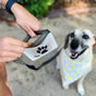 A person holds a treat near a clear (re)zip Take Me Too Pet Bag + Carabiner with a paw print design, while a happy leashed dog in a yellow and white bandana looks up expectantly beside the handy treat bag.