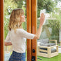 A woman smiles as she cleans a glass door with Reel Premium Bamboo Paper Towels (135 2-ply sheets per roll). She stands indoors, with an outdoor garden and furniture visible through the sparkling door.