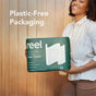 A smiling woman stands against a wooden wall, holding a pack of Reel Premium Bamboo Paper Towels—135 2-ply sheets per roll—in plastic-free packaging. White text reads “Plastic-Free Packaging.”.