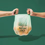 Two hands from opposite sides hold a semi-transparent Repurpose Compostable Small Bin Bag, Lavender Scented, with an orange object inside against a green background. The home-compostable bag reads “Replace it with Repurpose.”.