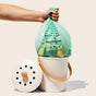 A hand holds a Repurpose Compostable Small Bin Bag, Lavender Scented, filled with food scraps above a white compost bin with wooden handle; the lid rests nearby against a plain light background.