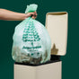 A hand holds a Repurpose Compostable Tall Kitchen Bag, Lavender Scented, over an open beige trash bin against a dark green background.