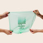 Four hands stretch a light green Repurpose Compostable Small Bin Bag with “Replace it with Repurpose. COMPOSTABLES” printed in black, set against a plain beige background.