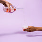 A hand pours a clear liquid from a decanter into Repurposes Stemless Cold Cup held by another hand. The soft purple background highlights the elegance and simplicity of this non-toxic gesture.