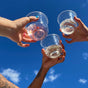 Three hands, displaying tattoos and rings with one featuring vivid blue nail polish, hold Repurposes Stemless Cold Cups filled with sustainable drinks. The scene shows two clear and one pink beverage raised towards a clear blue sky, celebrating individuality.