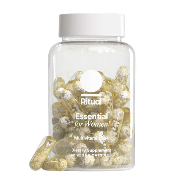A clear bottle labeled Ritual Women's Multivitamin 50+ holds yellow capsules with white bead-like spheres inside. Two capsules are placed in front of the upright bottle, ideal as a post-menopause multivitamin option.