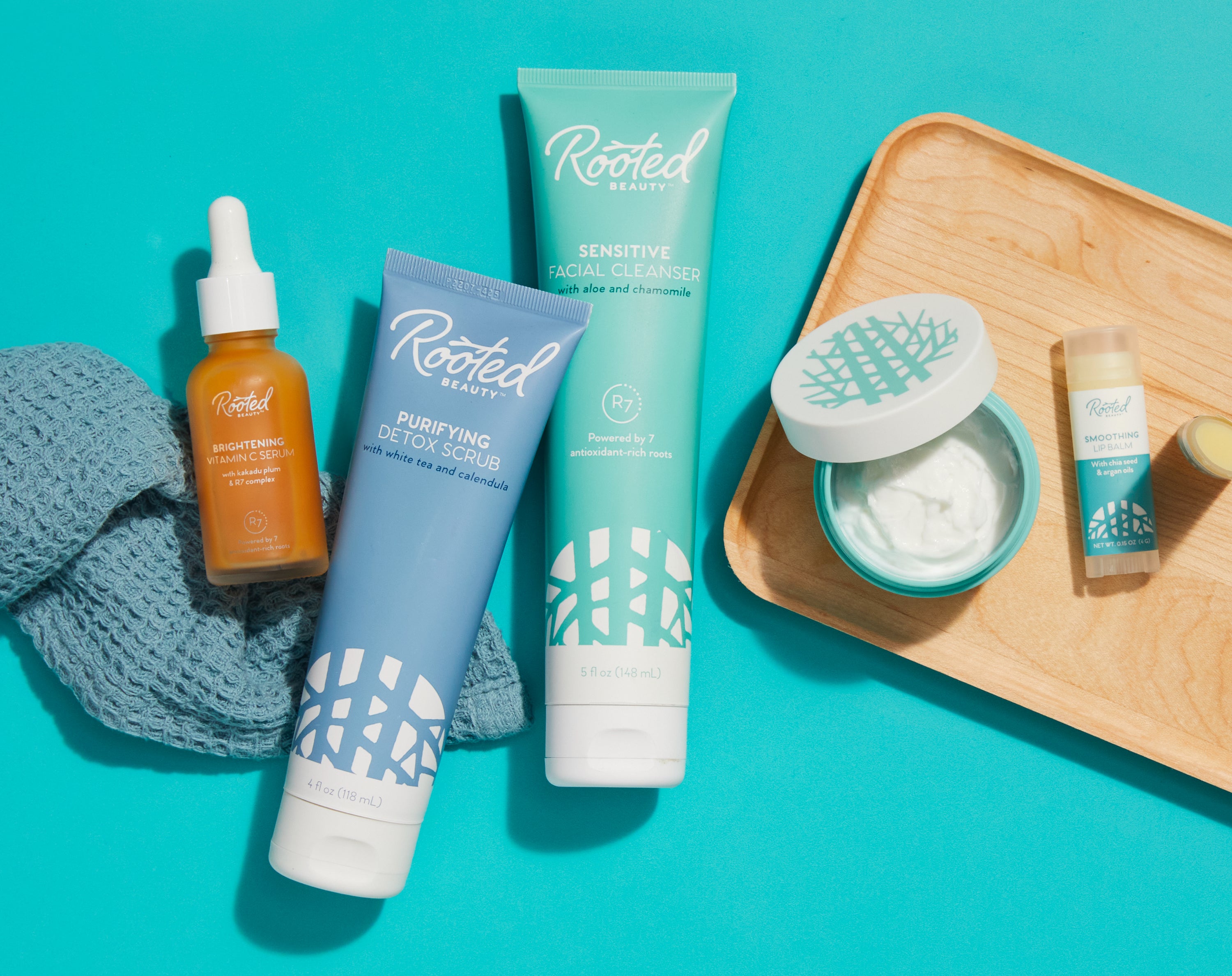 Image of a collection of products from Rooted beauty. Includes brightening vitamin C serum, purifying detox scrub, sensitive facial cleanser, and smoothing lip balm.