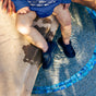 A toddler wearing an Eco Snap Swim Diaper by Green Sprouts, made from recycled polyester, sits happily by the pools edge in black water shoes and a blue fish-patterned outfit, splashing their feet while an adults hand rests on the blue tiled border.