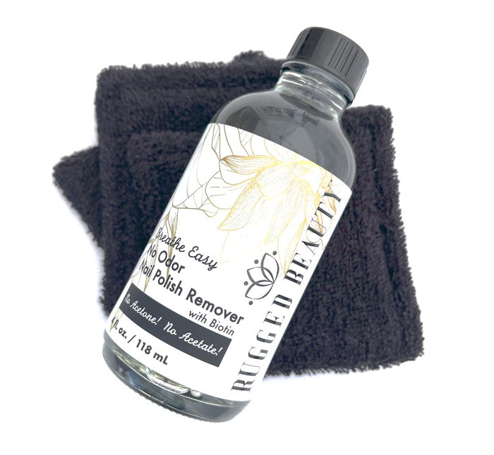 A clear bottle of Rugged Beauty Breathe Easy No Odor Nail Polish Remover rests on a folded black cloth, with a label displaying floral artwork and product details.