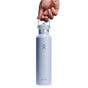 A hand holds the handle of a light blue Hydro Flask Standard Mouth Bottle with Flex Straw Cap, 24 oz, featuring the iconic logo and a silver cap. Its TempShield double-wall insulation keeps drinks at an ideal temperature, while its sleek design is highlighted against a white background.