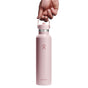 A hand holds a pastel pink Hydro Flask Standard Mouth Bottle with Flex Straw Cap, 24 oz. It has a cupholder-compatible design, TempShield double-wall insulation, and features a silver lid with a minimalistic logo on top, set against a plain white background.