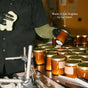 A worker wearing gloves inspects Seasonal Mini Candle Gift Set soy wax candles in amber glass with gold lids on an assembly line, with text above reading Made in Los Angeles by our team.