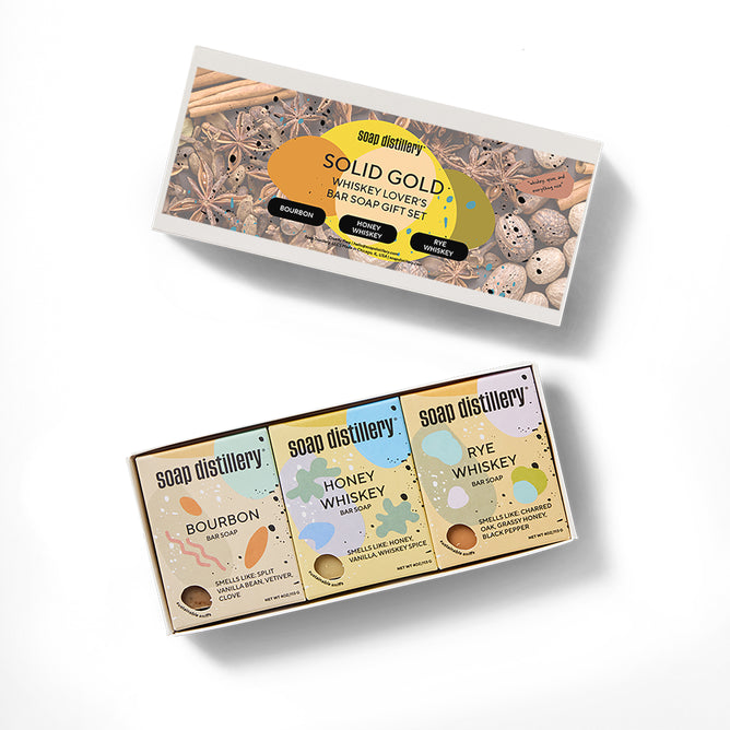 The Solid Gold Three Soap Gift Set by Soap Distillery features Bourbon, Honey Whiskey, and Rye Whiskey soaps in a display box with spice illustrations—an ideal whiskey-inspired gift for any enthusiast.