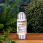 A silver aluminum bottle labeled Soap Distillery Hand + Body Wash rests on a wooden surface among green plants and blurred foliage, evoking the fresh essence of this scented wash.