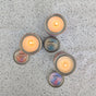 Five Soap Distillery Soy Wax Candles in metal tins sit on a speckled surface. Visible labels: “PINK GRAPEFRUIT LAVENDER,” “SAGE + SALTED PELT,” and “SUNSET FIG.” These paraffin-free candles offer a cozy, inviting glow.
