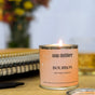 A Soap Distillery Soy Wax Candle sits on a surface beside stacked notebooks and a pen, with colorful flowers softly blurred in the background.