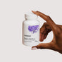 A hand with manicured nails holds a white bottle labeled "Thorne Women's Libido Boost Capsules," featuring a purple abstract design, against a plain light background.